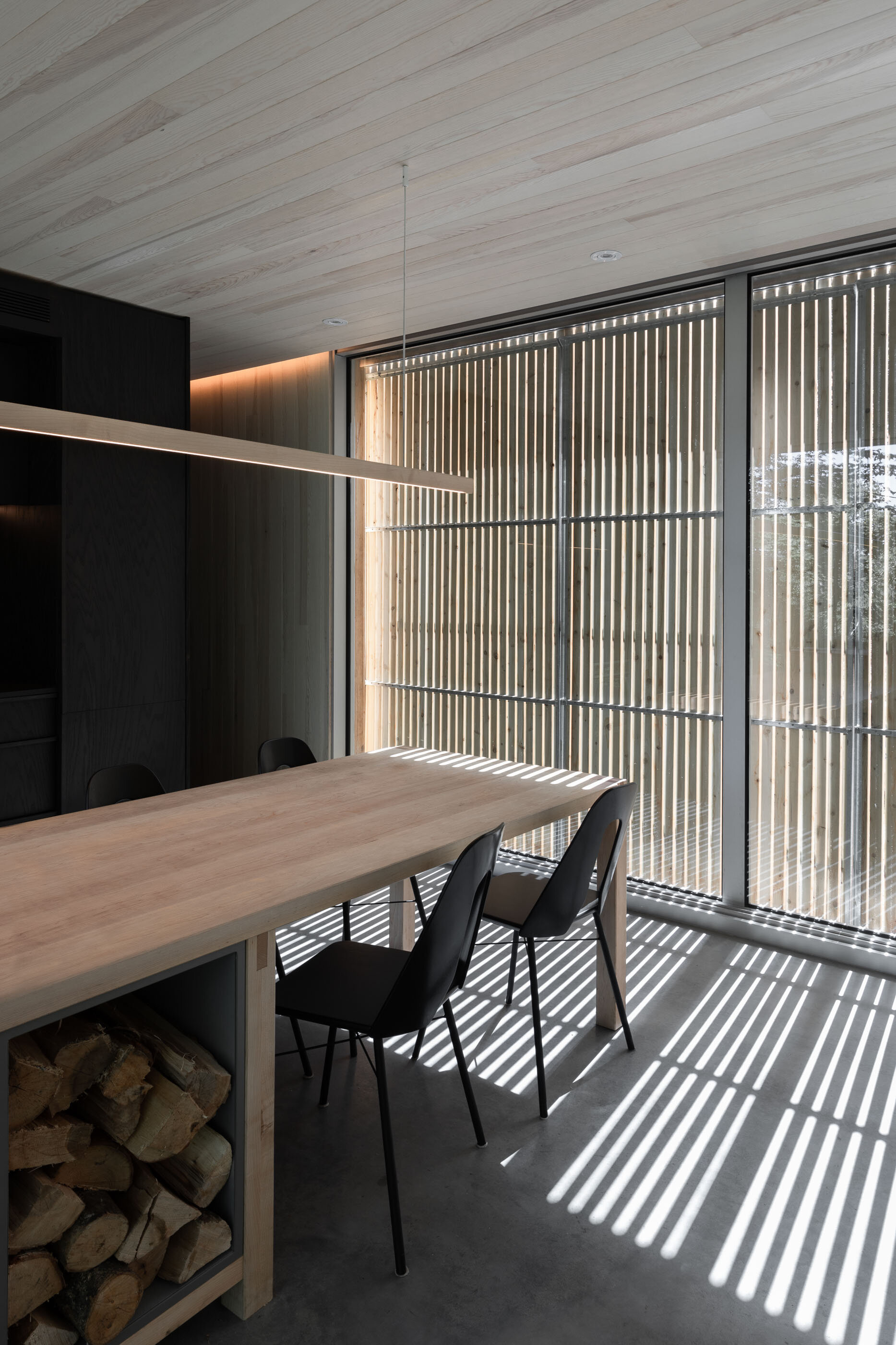 Modern dining area with a wooden table, black chairs, large window with wooden slats creating striped shadows, and wood storage with logs underneath.