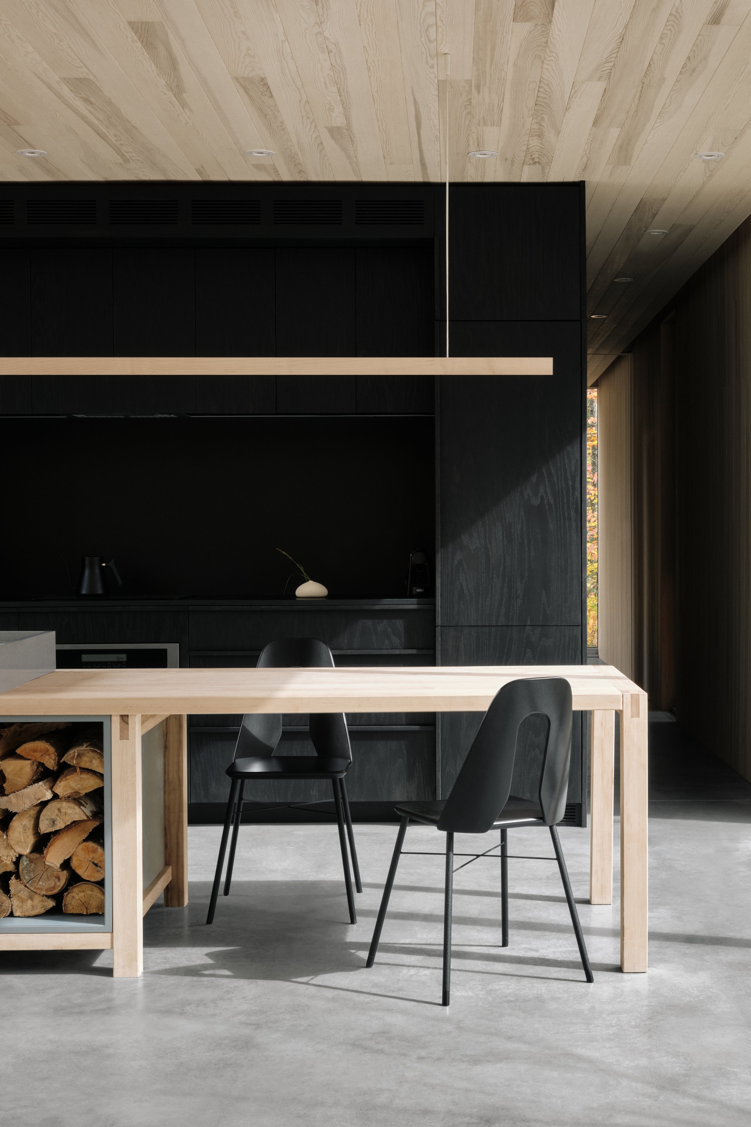 Minimalist modern kitchen with black cabinetry, light wooden accents, a wooden dining table, and black chairs, with a wood storage nook and a small potted plant.