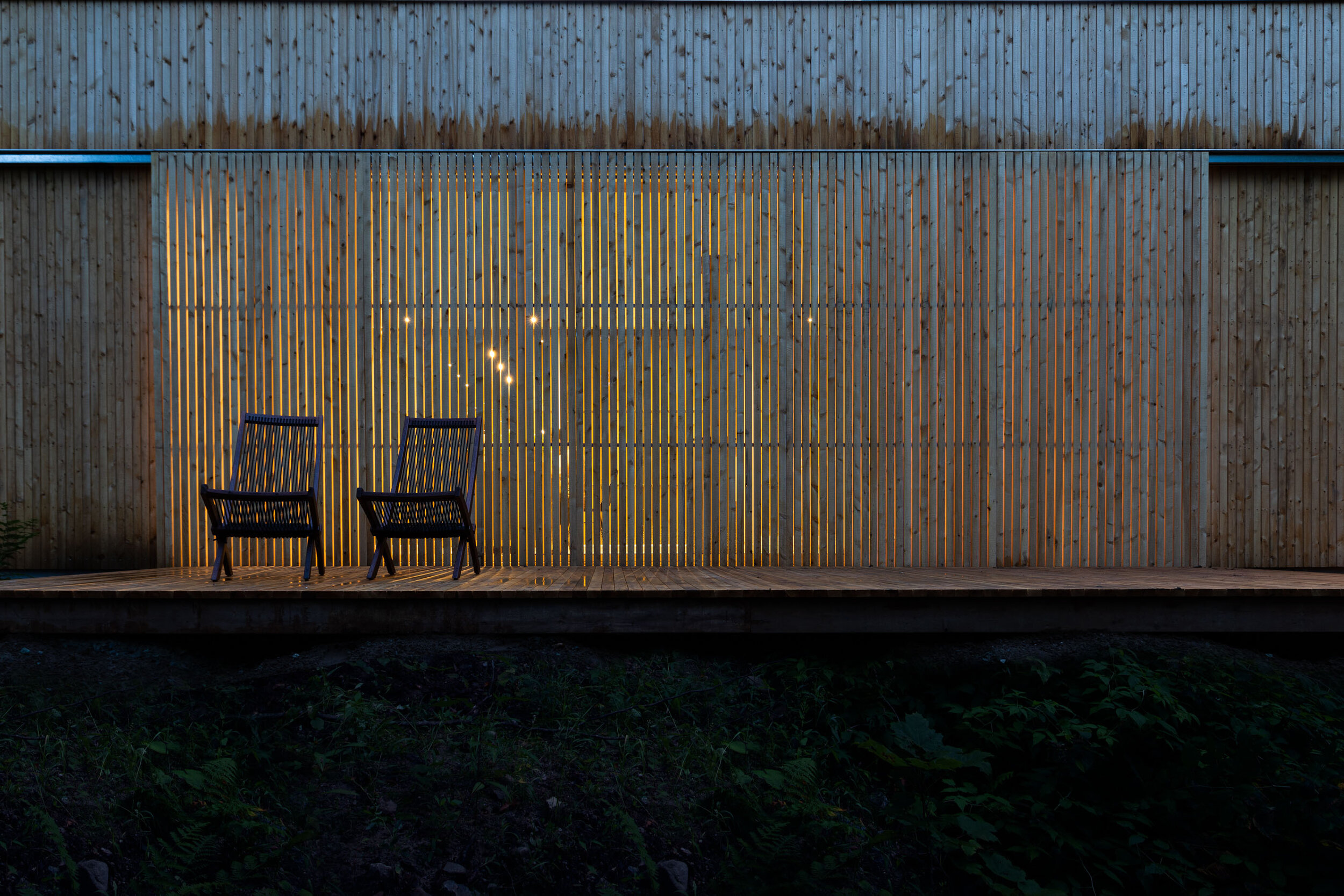 Two black chairs on a wooden deck in front of a wooden slat fence at dusk, with lights visible through the fence.