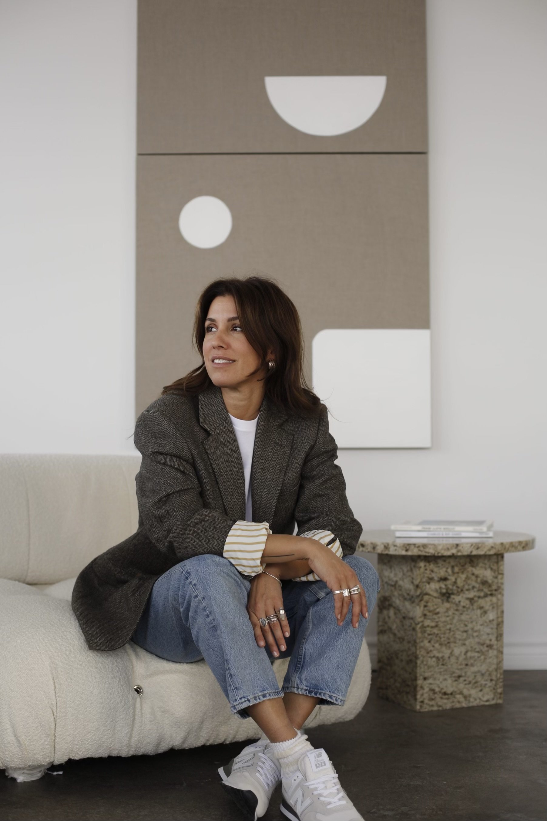 A woman sitting on a light-colored sofa in a modern room with abstract wall art, wearing a blazer, jeans, and sneakers, looking to the side.
