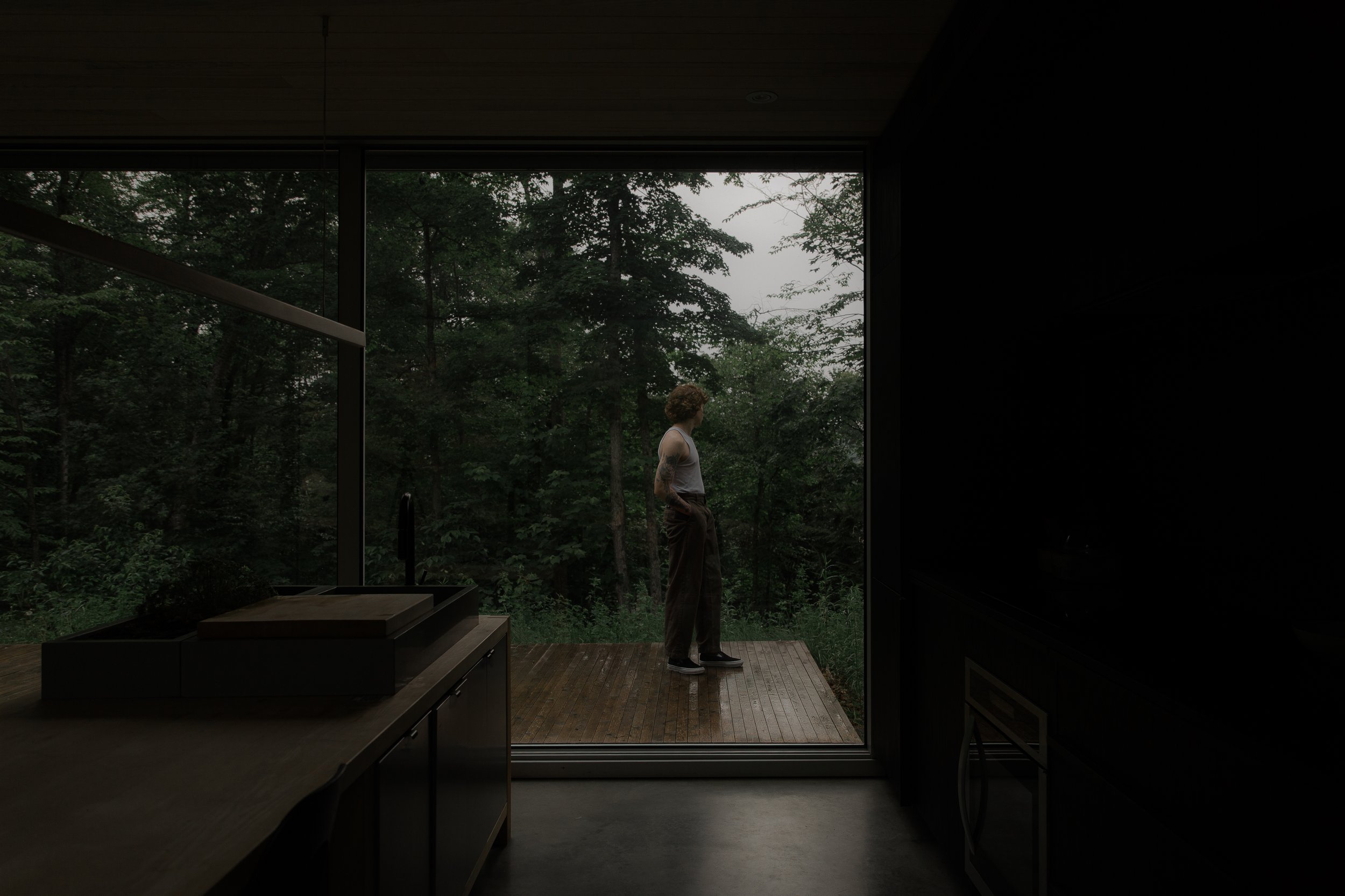 A person with curly hair, wearing a sleeveless shirt and loose pants, standing on a wooden deck outside a modern glass house surrounded by dense green trees.
