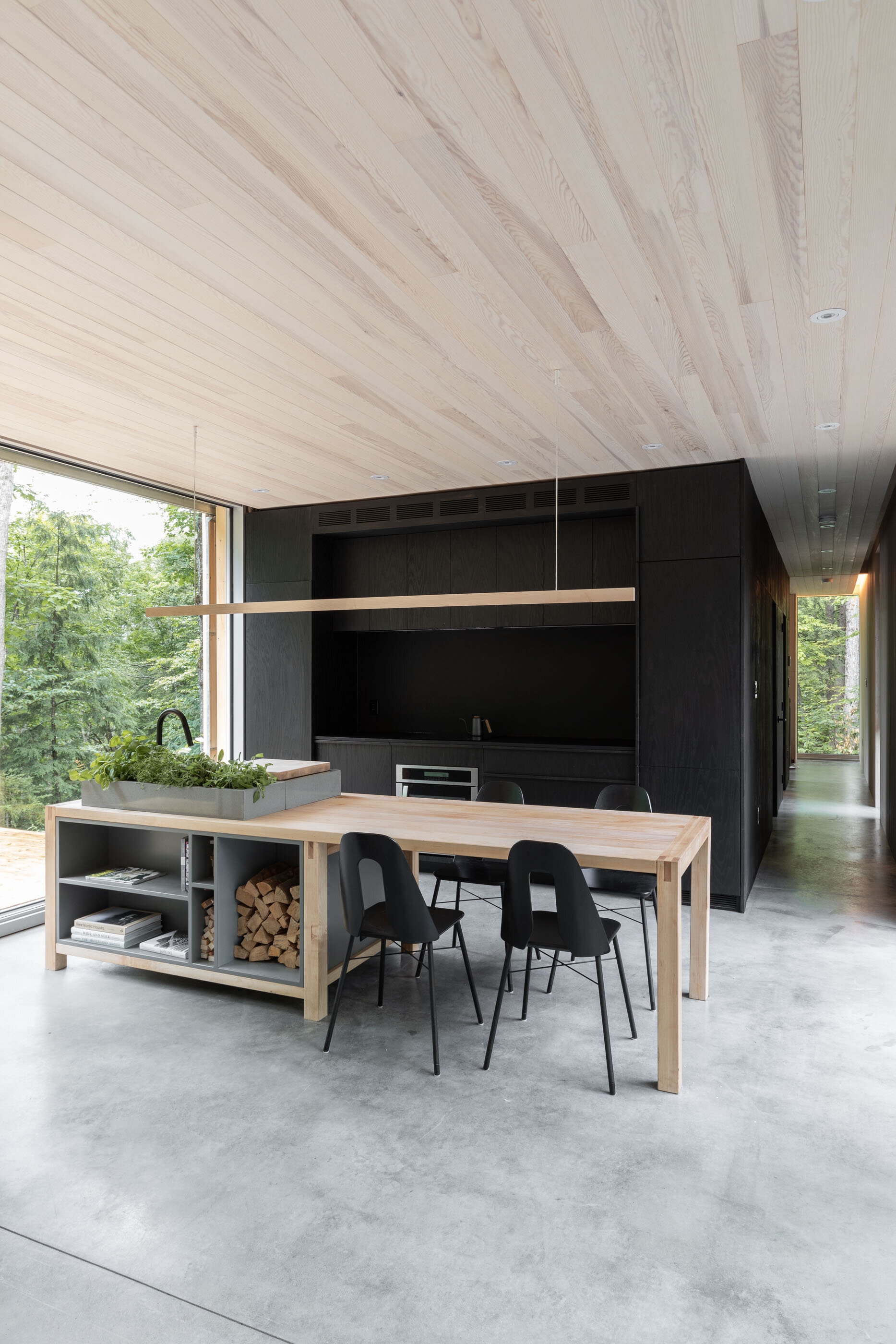 Modern dining area with light wood table, black chairs, and built-in storage with firewood, in front of large window showing greenery.