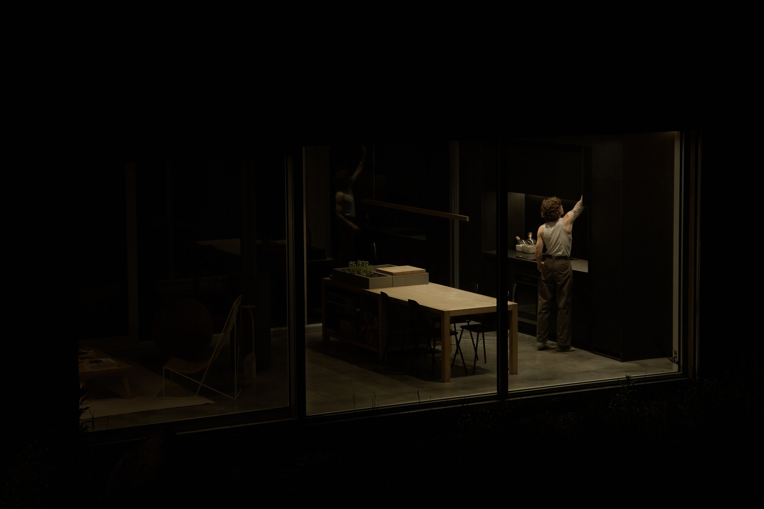 A person with curly hair in a sleeveless shirt standing at a kitchen counter, with another person reflected in a mirror, inside a modern, darkly lit room with a dining table and chairs visible.