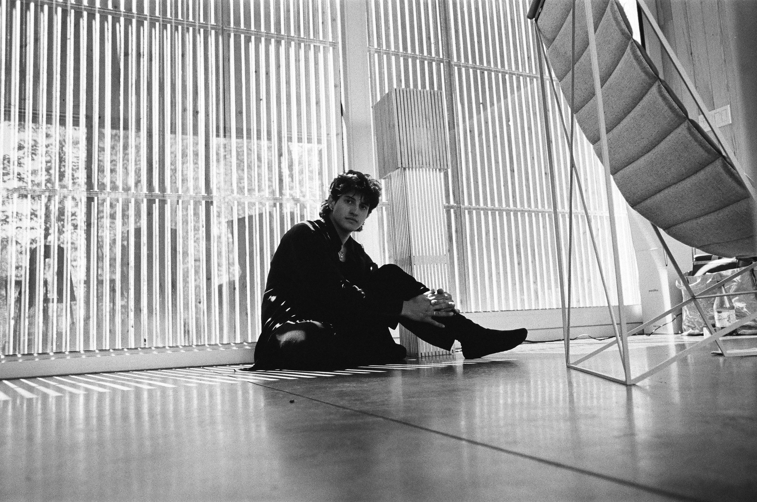A young person with dark hair, sitting on the floor of a modern interior with large windows and vertical blinds, wearing dark clothing and looking towards the camera.