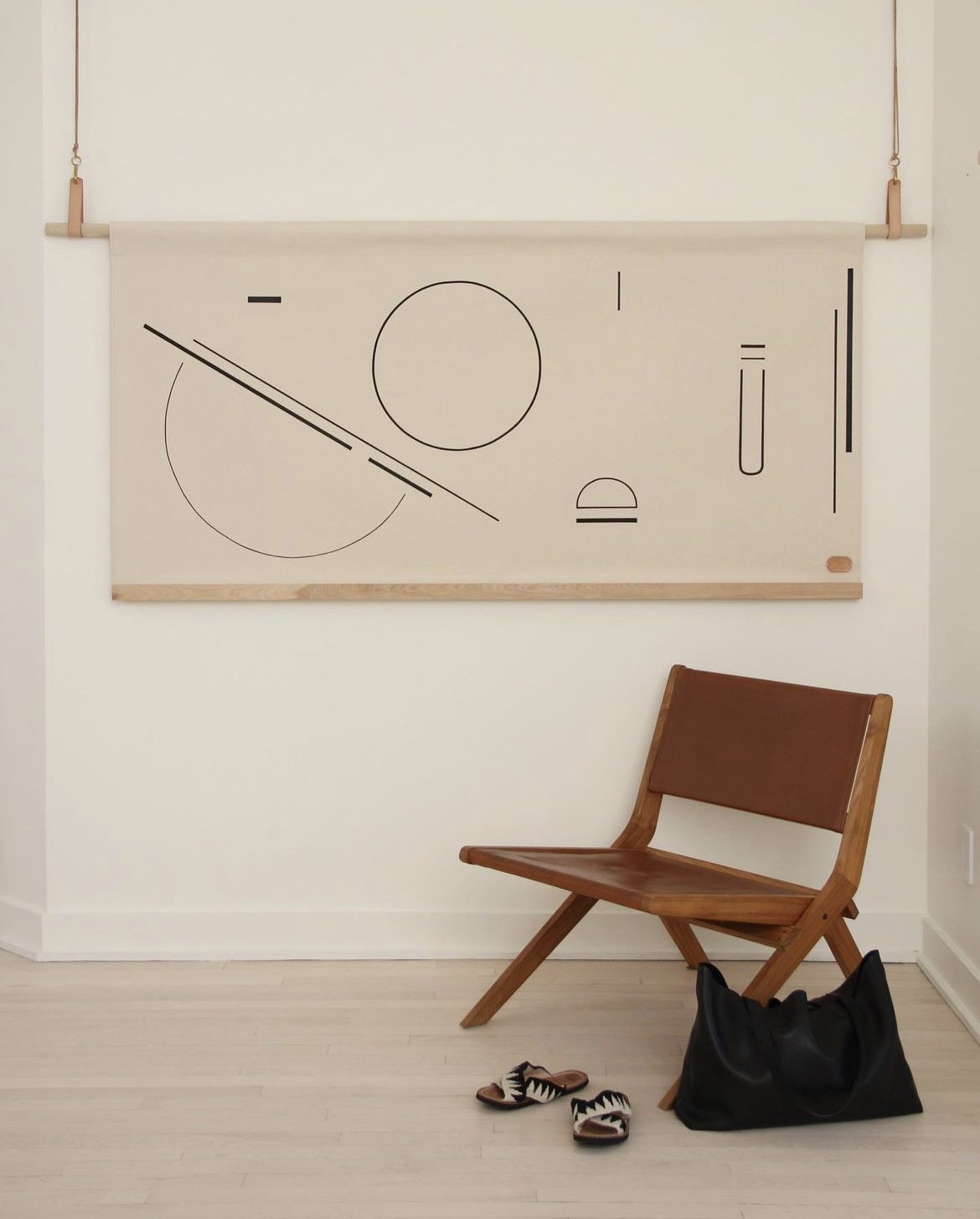 A modern interior with a wooden chair, a black bag, a pair of shoes with animal print straps, and an abstract black line drawing on a large wall hanging.