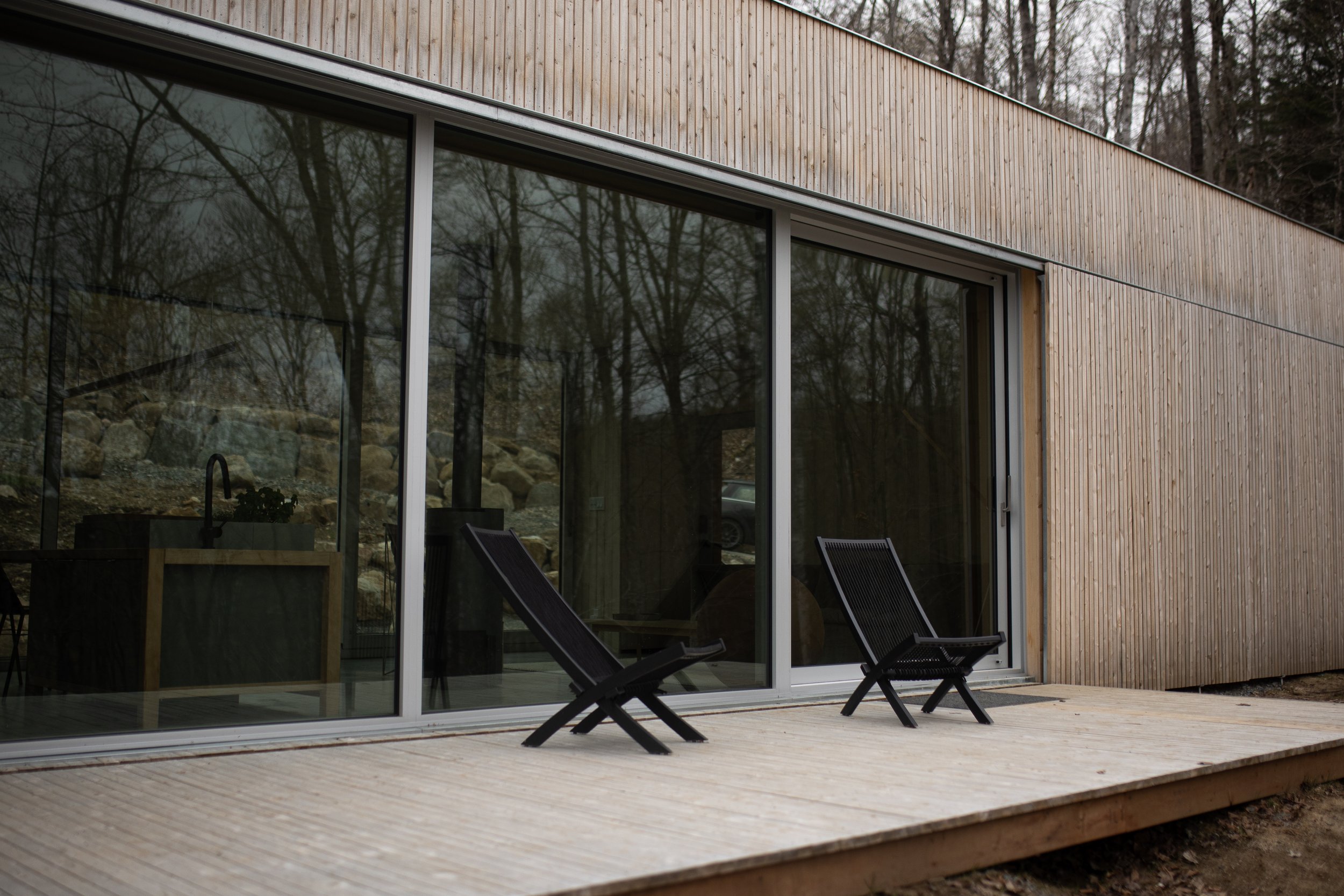 Modern house with wooden exterior, large glass sliding doors, and a wooden deck with two black lounge chairs outside.