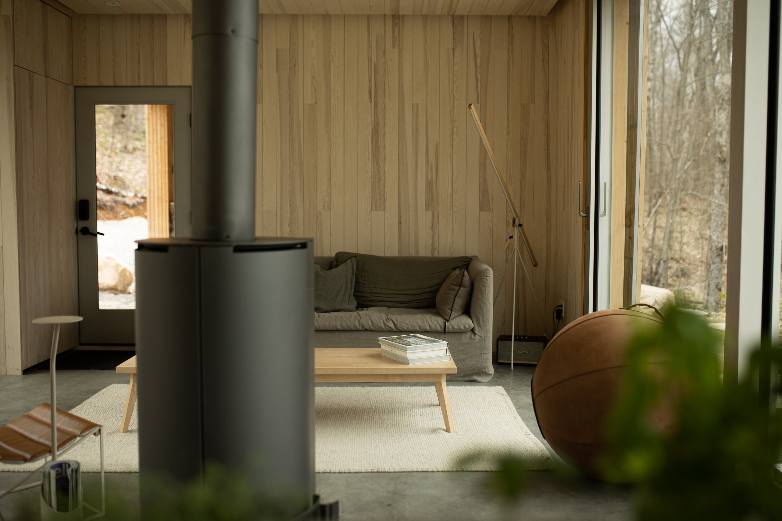 Living room with a beige sofa, wooden coffee table with magazines, large windows, and a view of trees outside.