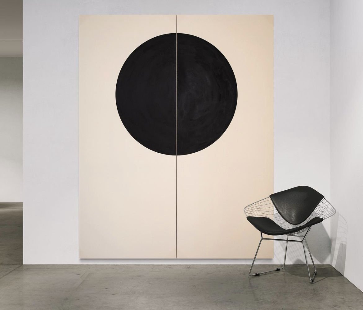 A modern black circle artwork on beige divided canvas wall behind a black chair with a wire frame.
