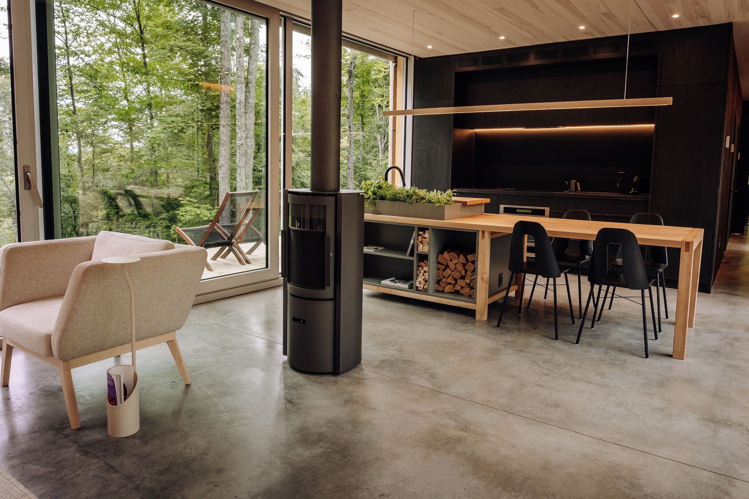 Modern open-concept living and dining area with large floor-to-ceiling windows overlooking a green forest, featuring a beige armchair, a small side table, a wood stove, and a wooden dining table with black chairs, all with minimalistic decor and conc