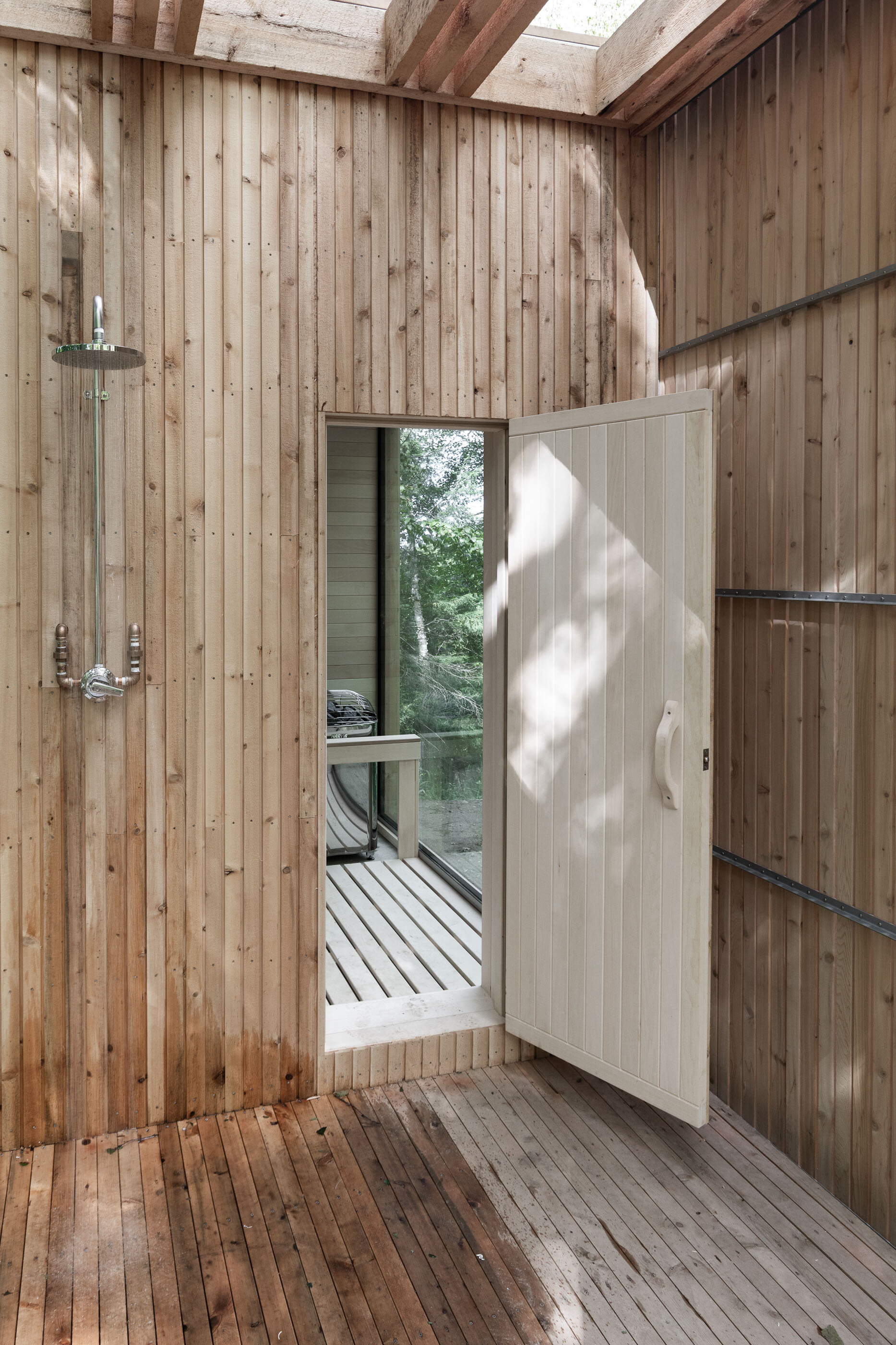 Interior of a wooden outdoor shower with a glass door leading to a small patio or balcony surrounded by trees.