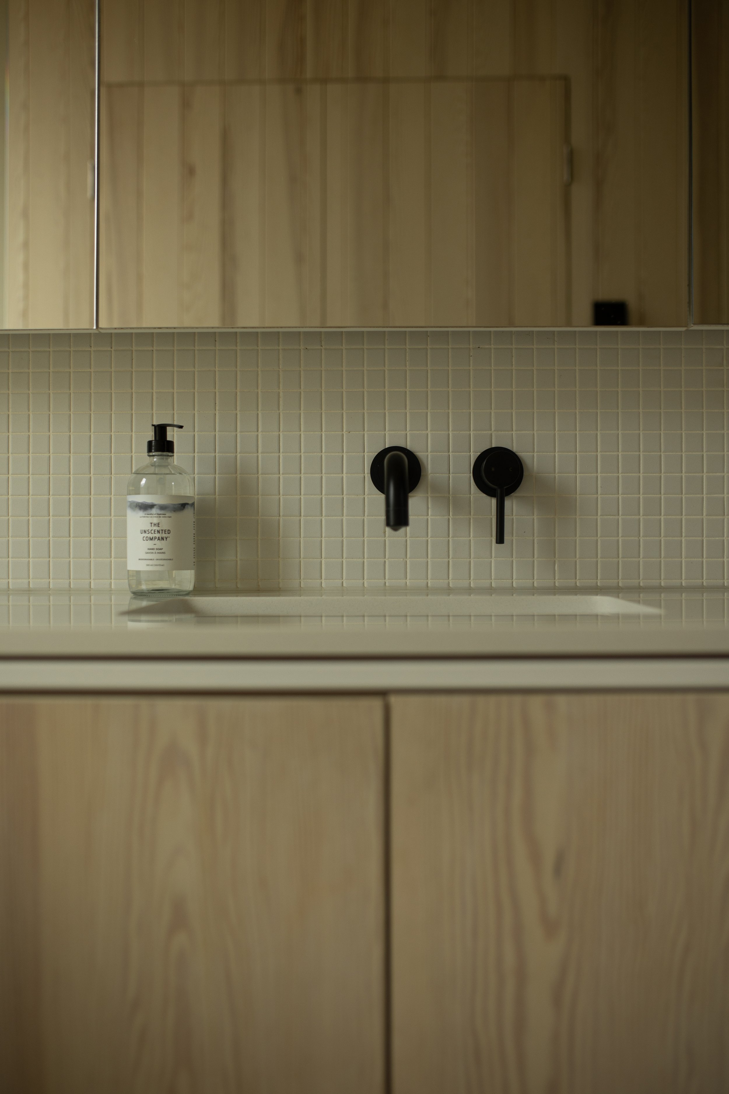 A bathroom sink area with a soap dispenser on the left, a beige tiled backsplash, and black faucet and control knobs. There is a wooden cabinet below and a mirror above.