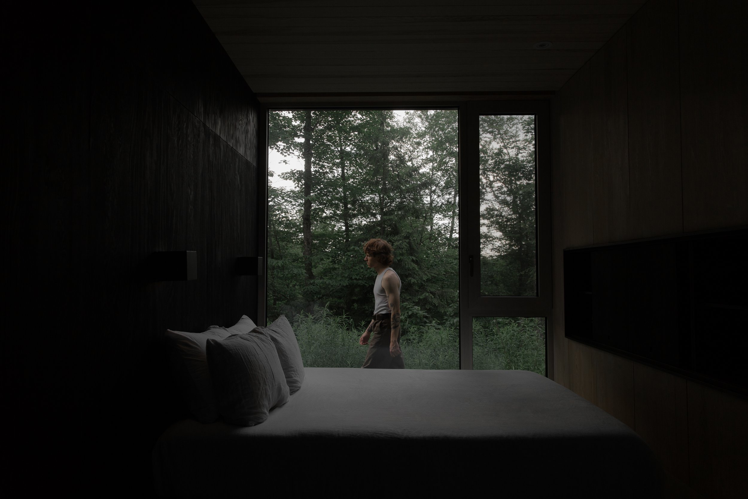 View of a bedroom with a large window showing a person walking outside in a lush green forest, inside the room there is a bed with pillows.