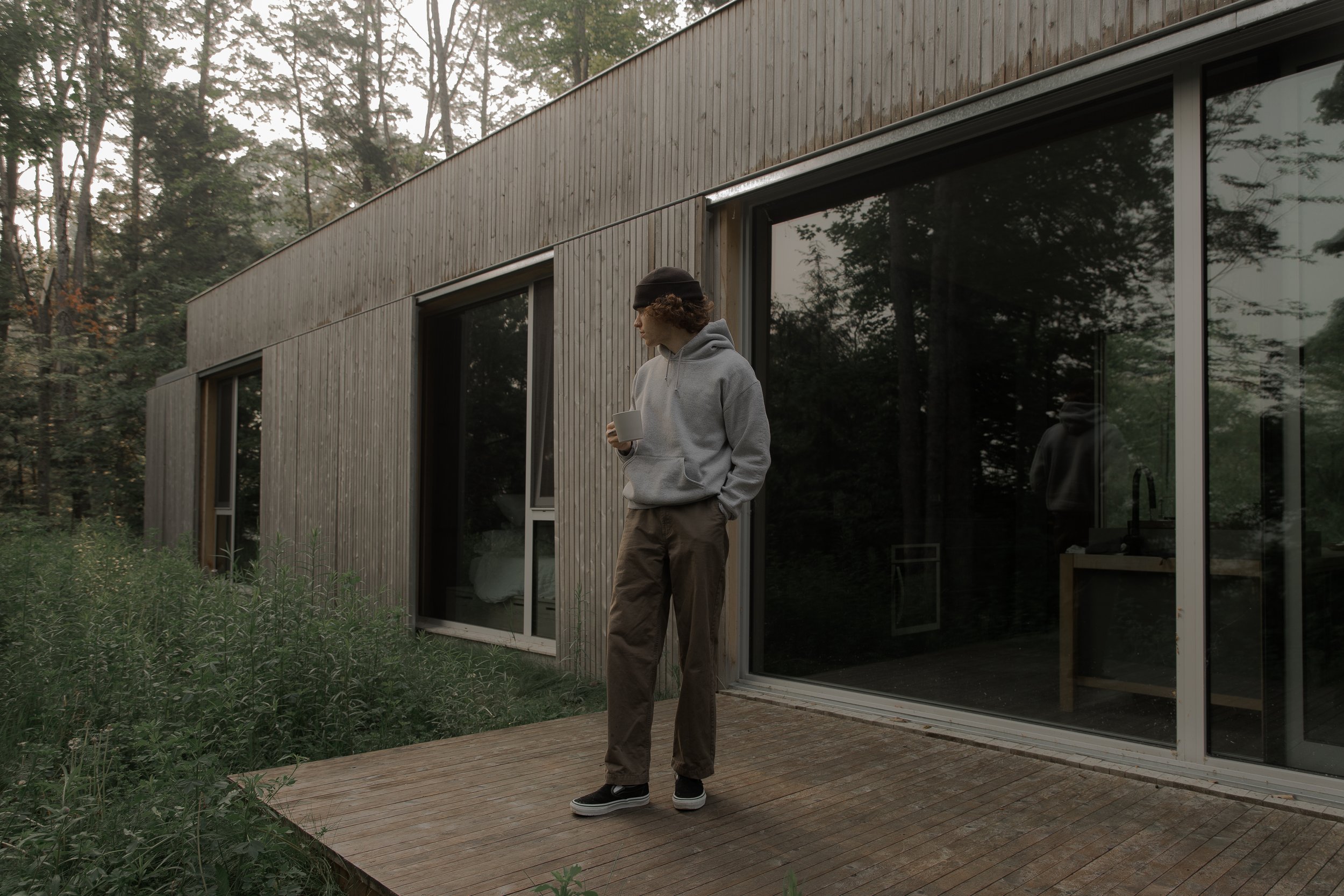 A young man standing outdoors on a wooden deck, holding a mug, near a modern house with large glass windows, surrounded by trees.