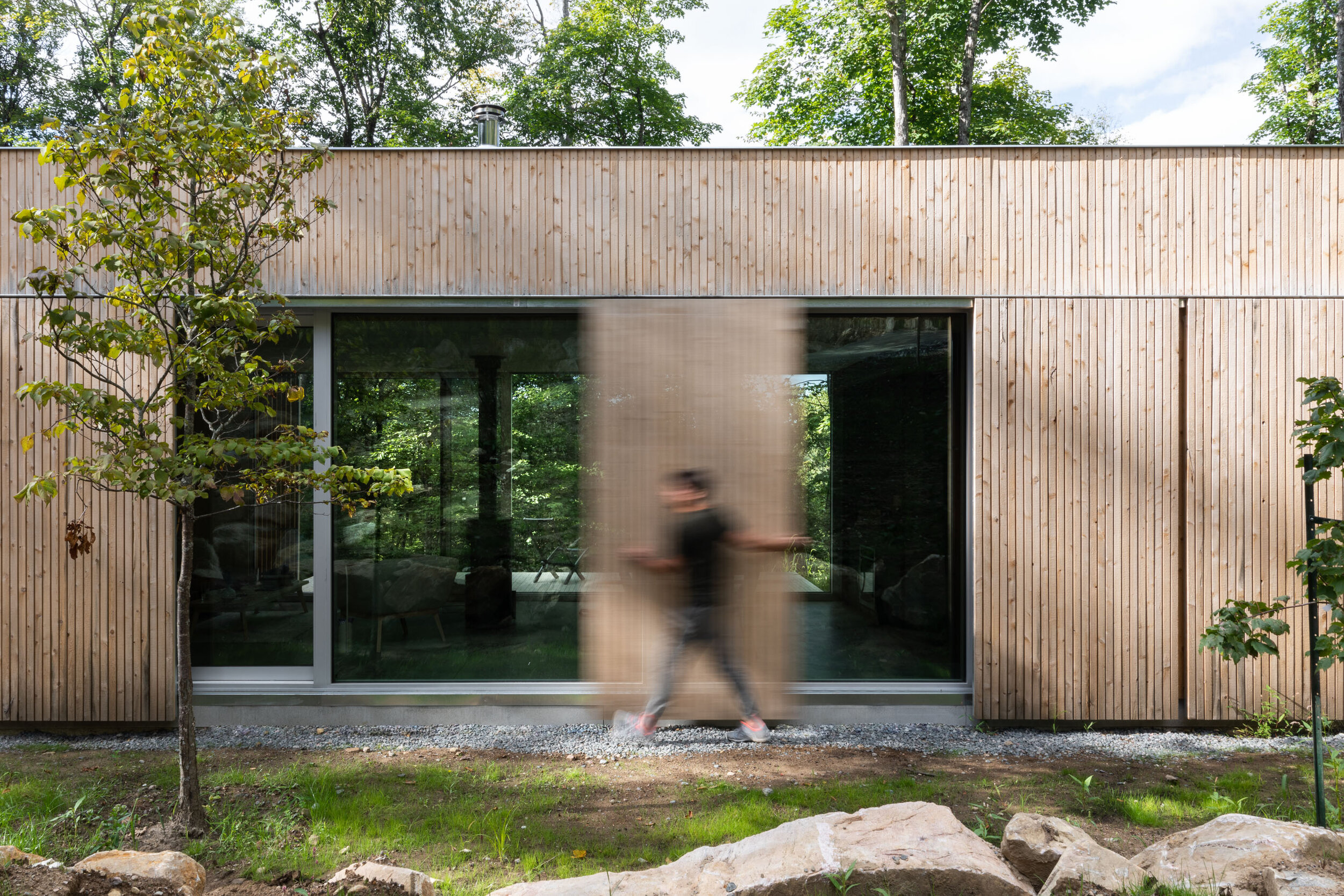 A man walking in front of a modern house with wooden exterior and large glass sliding door, with greenery and trees in the background.