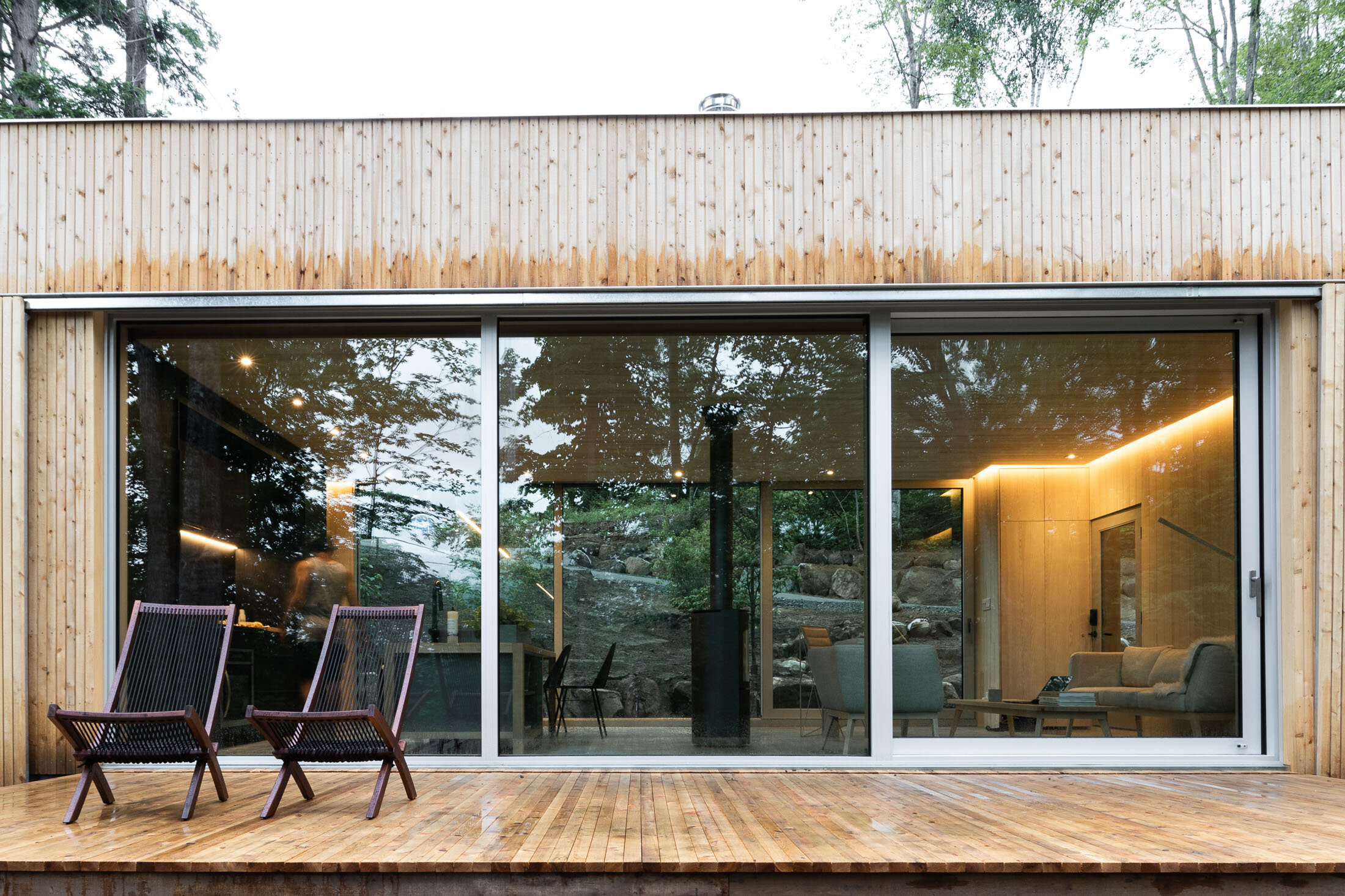 Modern house with large glass sliding doors, wooden exterior, and a wooden deck with two lounge chairs outside.