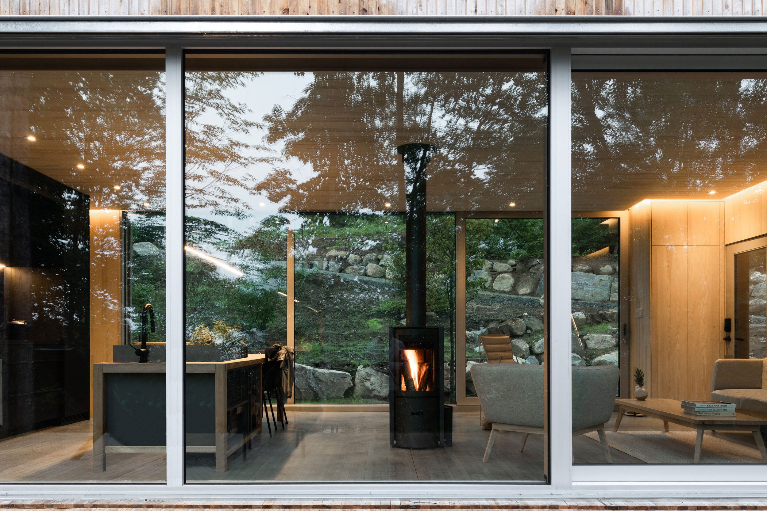 Living room with large glass windows, wood-paneled walls, and a wood stove in the center. Furniture includes a beige armchair, a gray armchair, and a coffee table with books and a small plant. Outside, rocks and greenery are visible through the windo