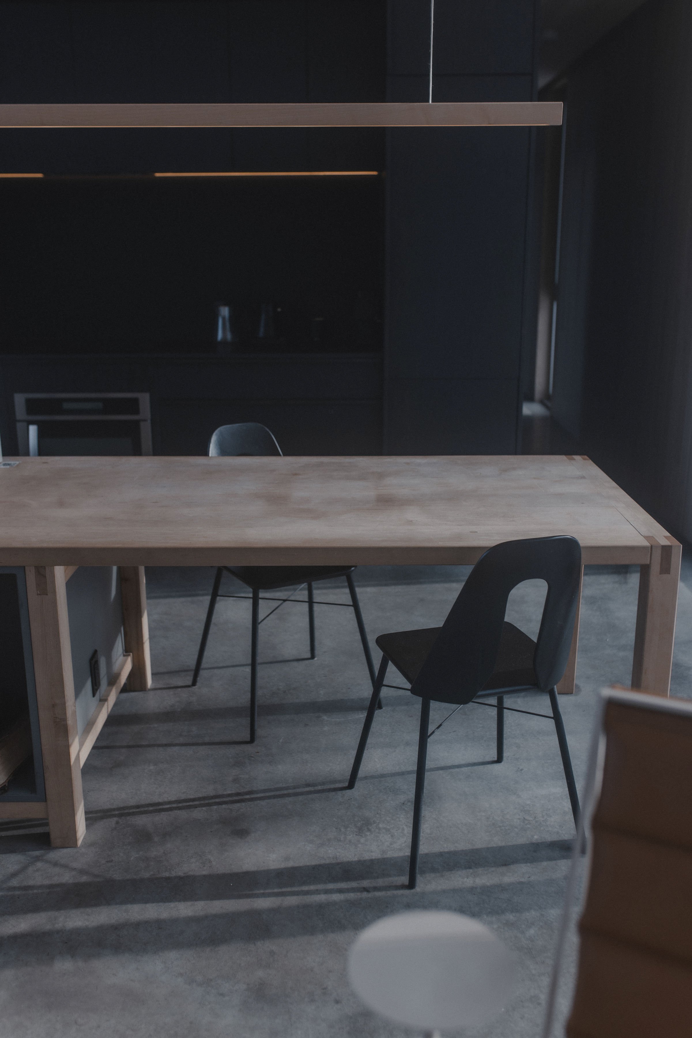 A minimalist office space with a wooden desk and three black chairs, set against dark cabinetry and a concrete floor.