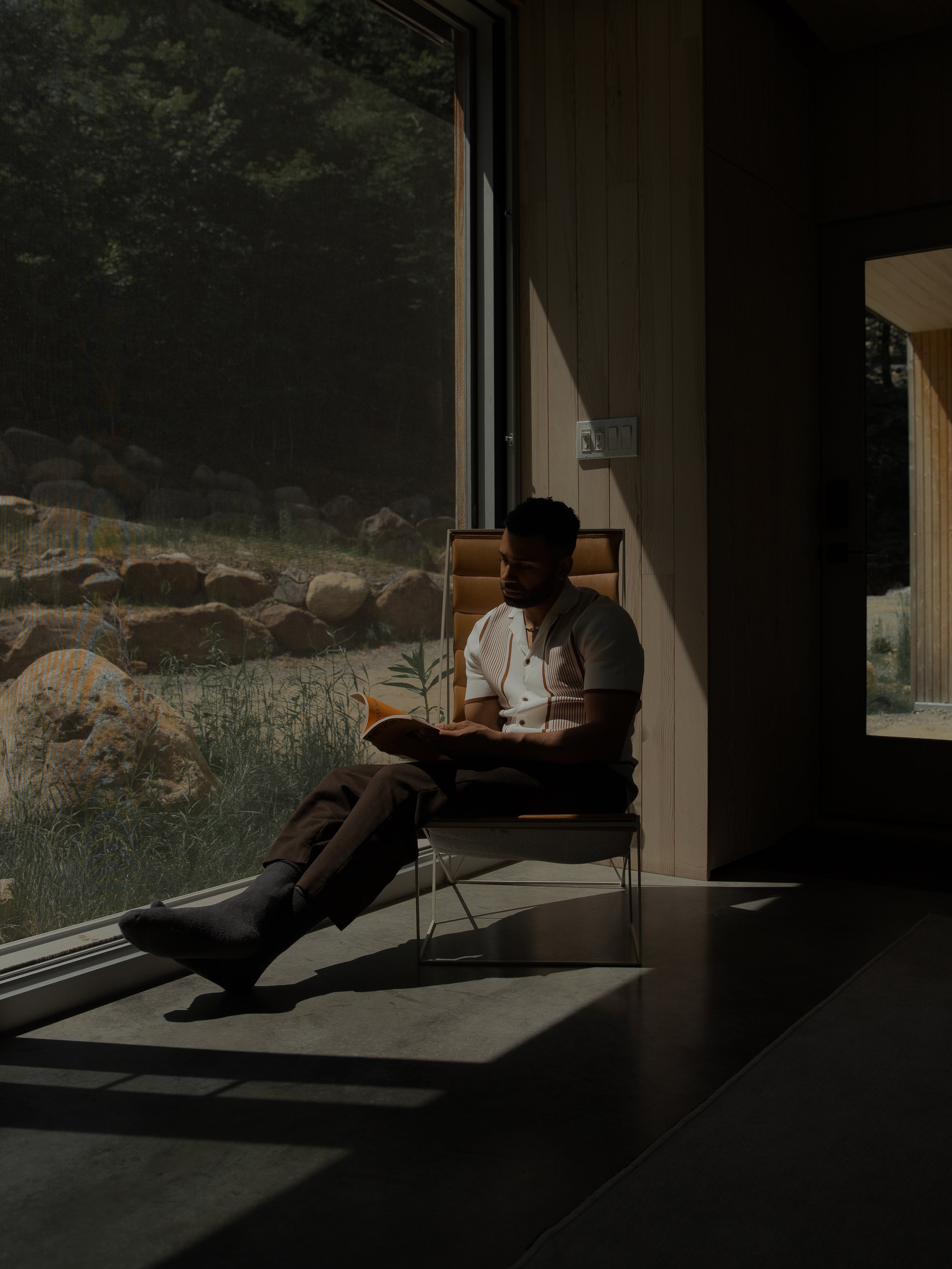 Man sitting in a chair by a large window, reading a book, with sunlight casting shadows inside a modern home.