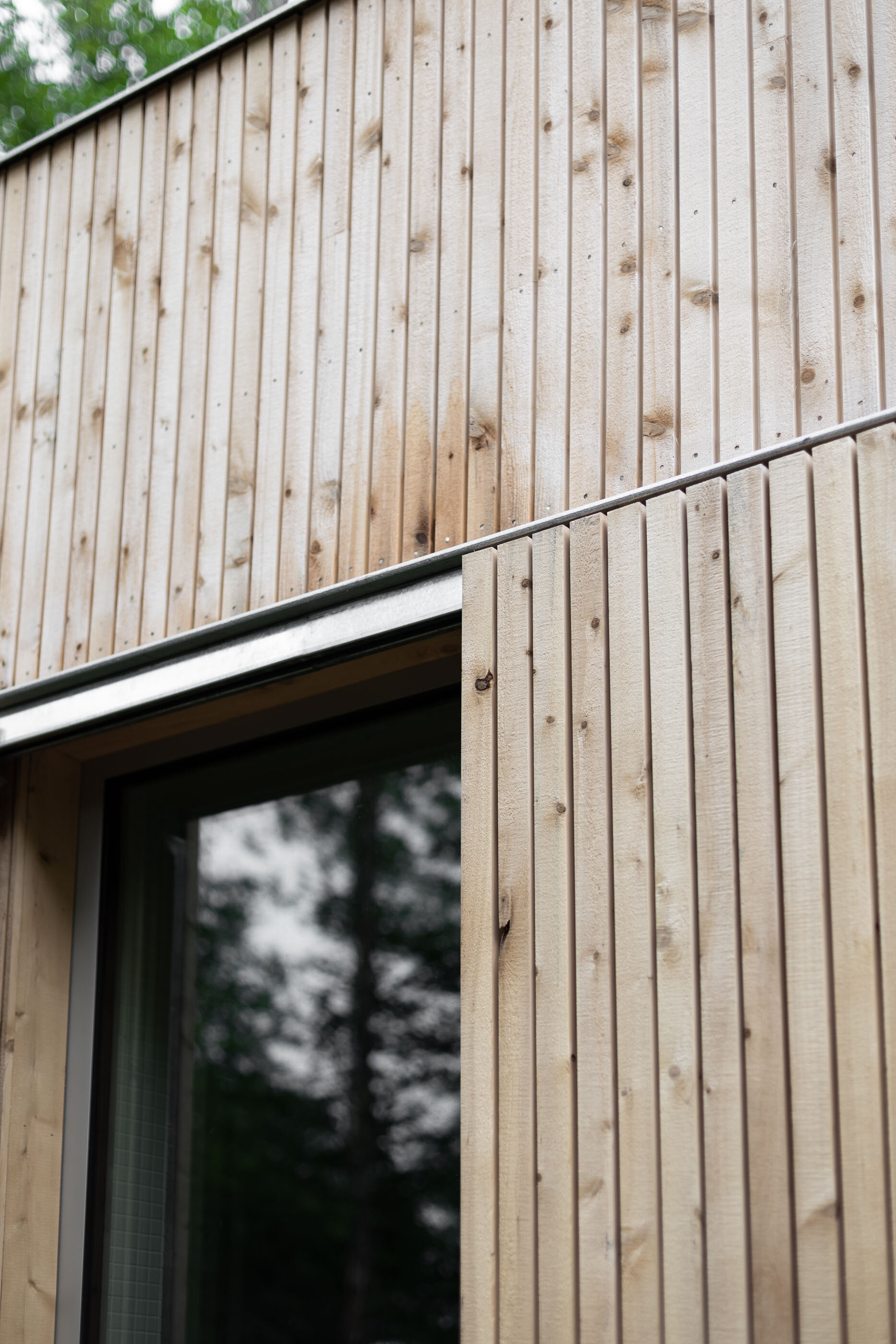 Close-up of a modern building exterior with vertical wood siding and a window, with trees reflected in the glass.