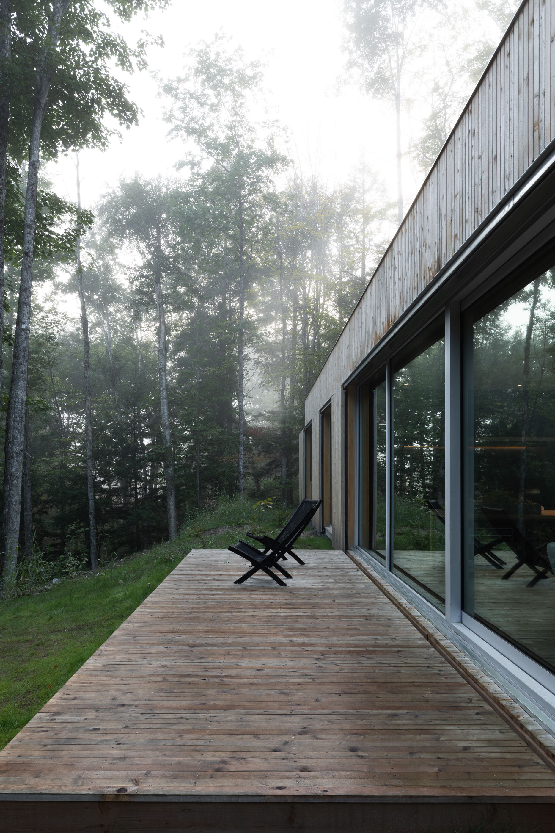A wooden patio with two black chairs overlooking a misty forest outside a modern house with large glass windows.