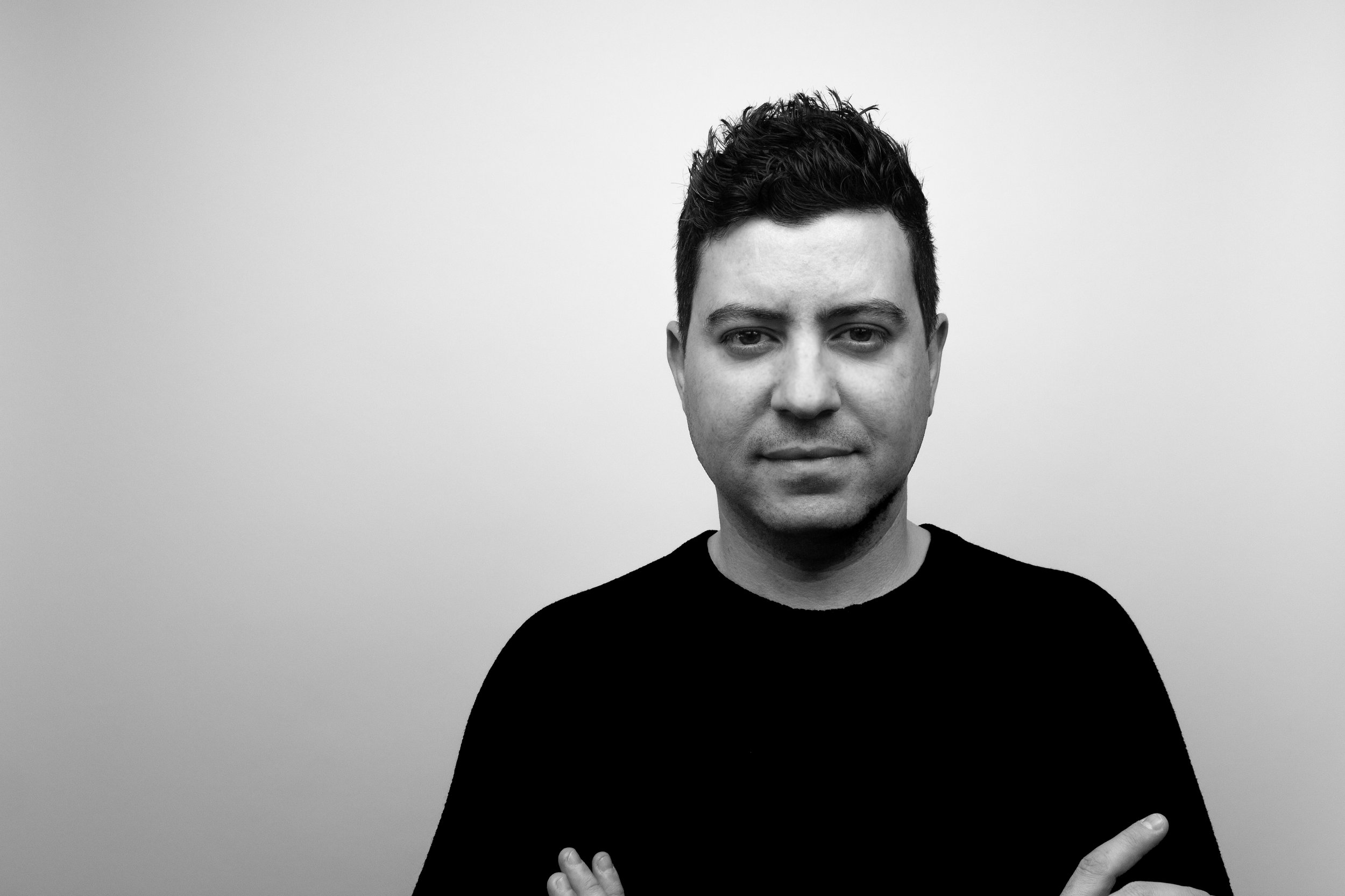 Black and white portrait of a young man with short, dark hair, crossing his arms, standing against a plain light-colored wall.