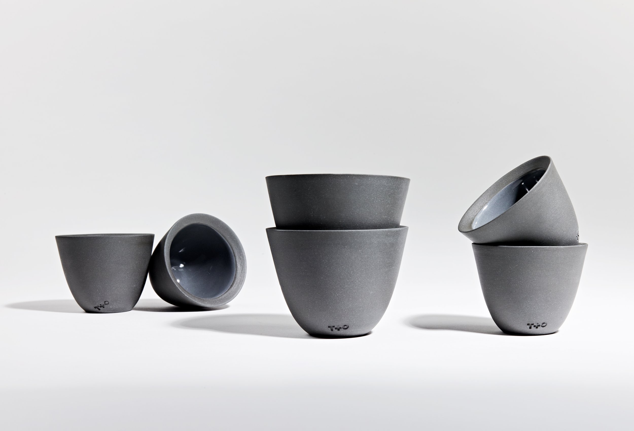 Six stacked gray ceramic bowls with black interior and the word 'TWO' engraved on the bottom, arranged in two groups on a white surface and background.