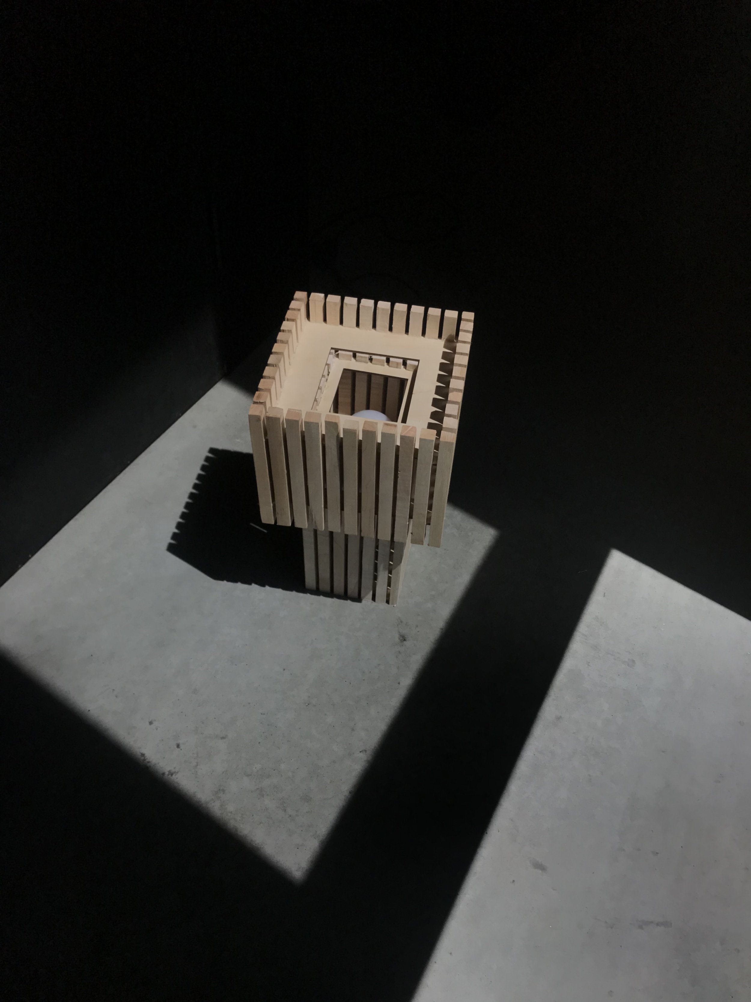 A rectangular wooden lamp made of evenly spaced vertical slats, casting shadows on the ground in a high-contrast setting with dark walls.