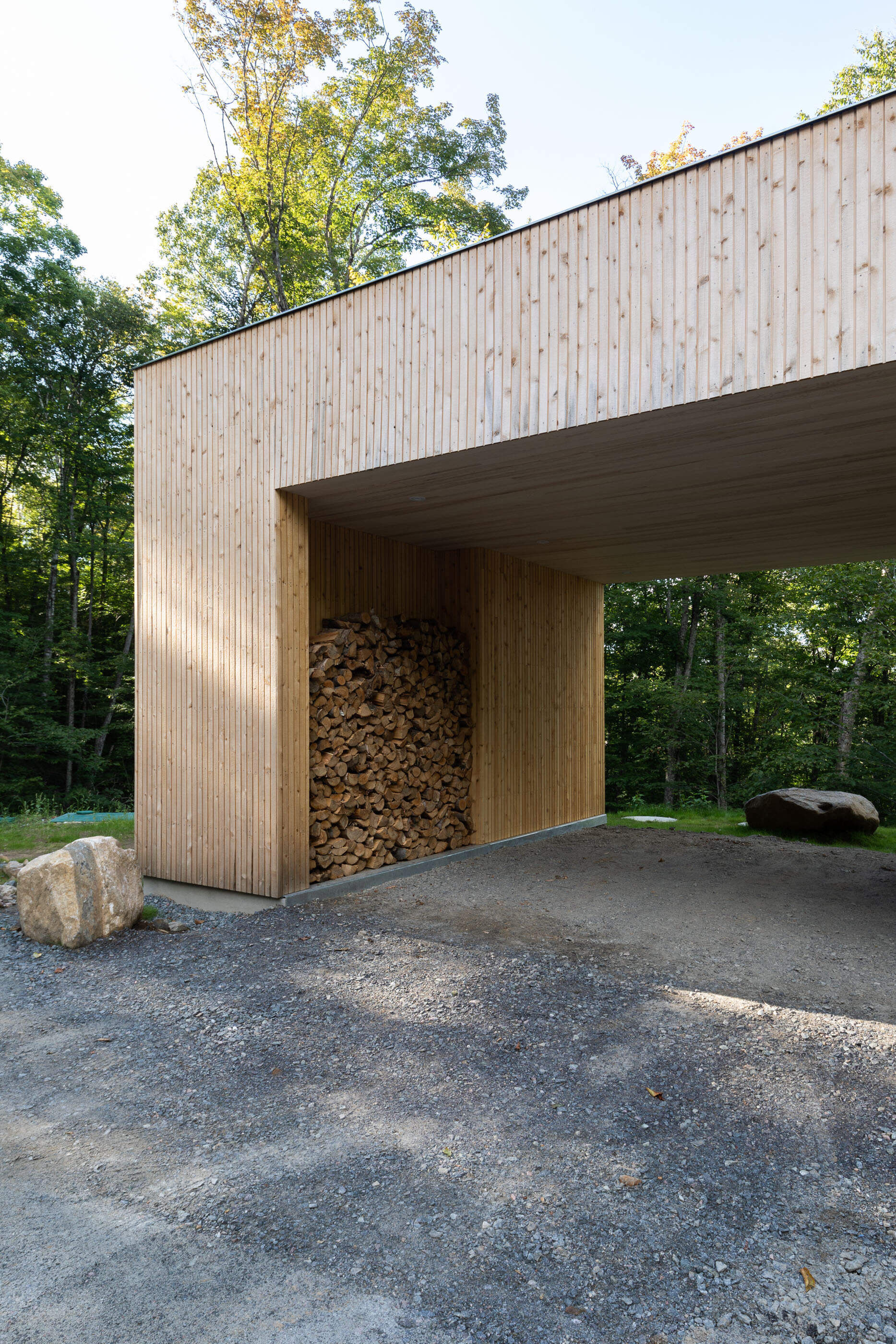 Modern wooden structure with a large stack of firewood stored underneath an overhang, surrounded by trees and a gravel driveway.
