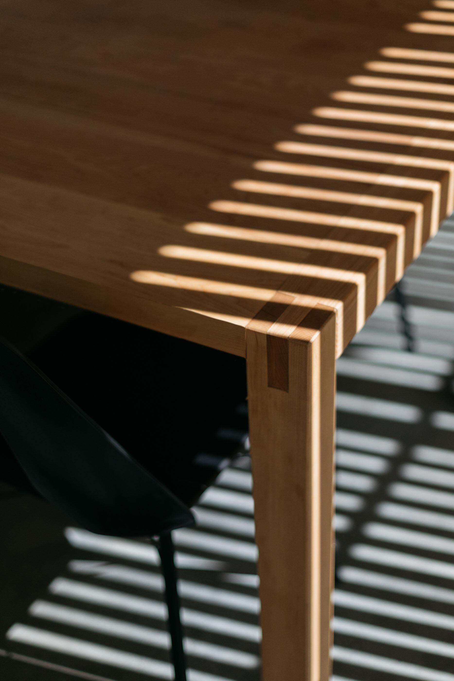 Close-up of a wooden table with slatted wood shadows cast on the surface and a chair with a dark seat in the background.