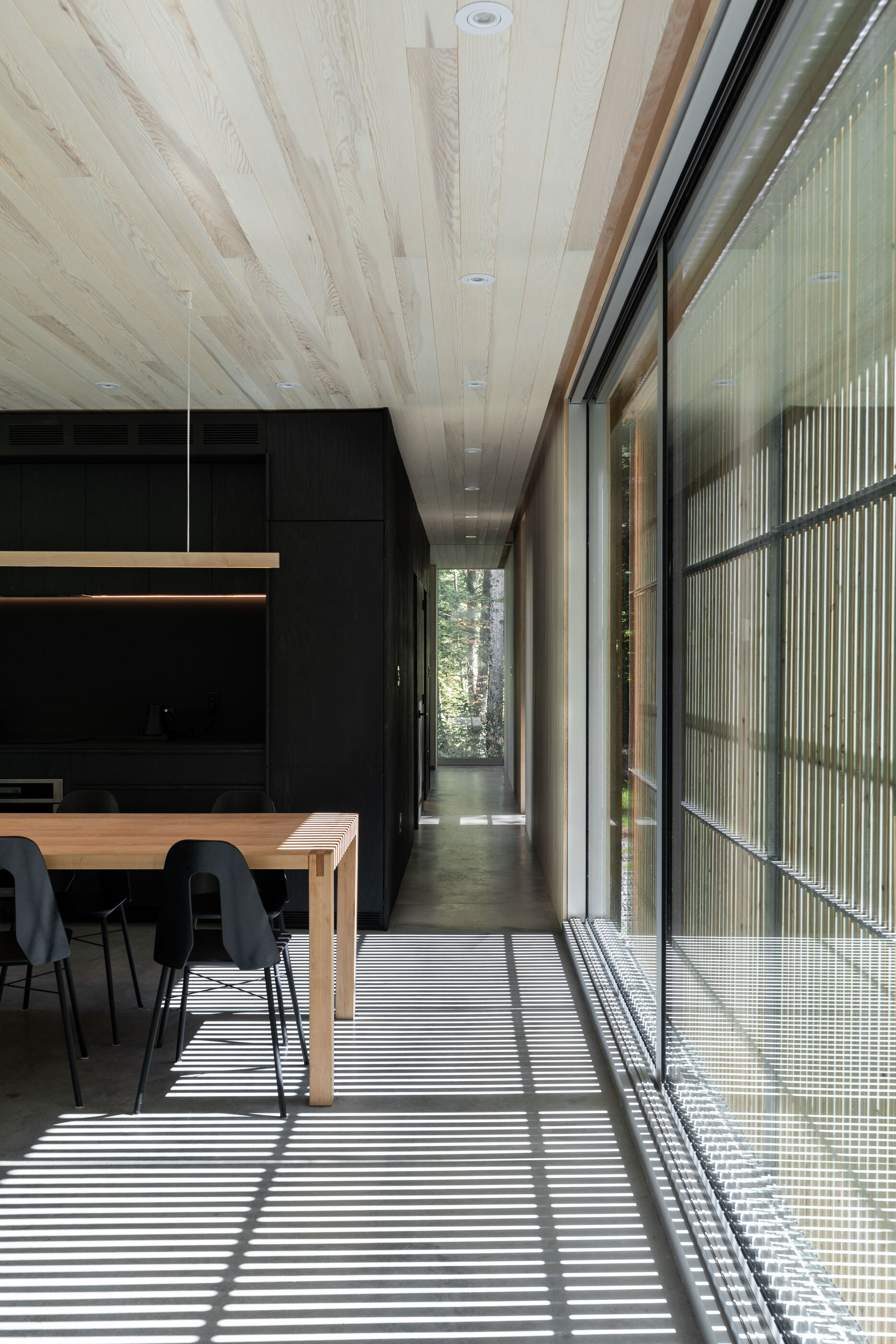 Modern dining room with a wooden table, black chairs, black walls, and large glass sliding doors casting striped shadows on the floor.