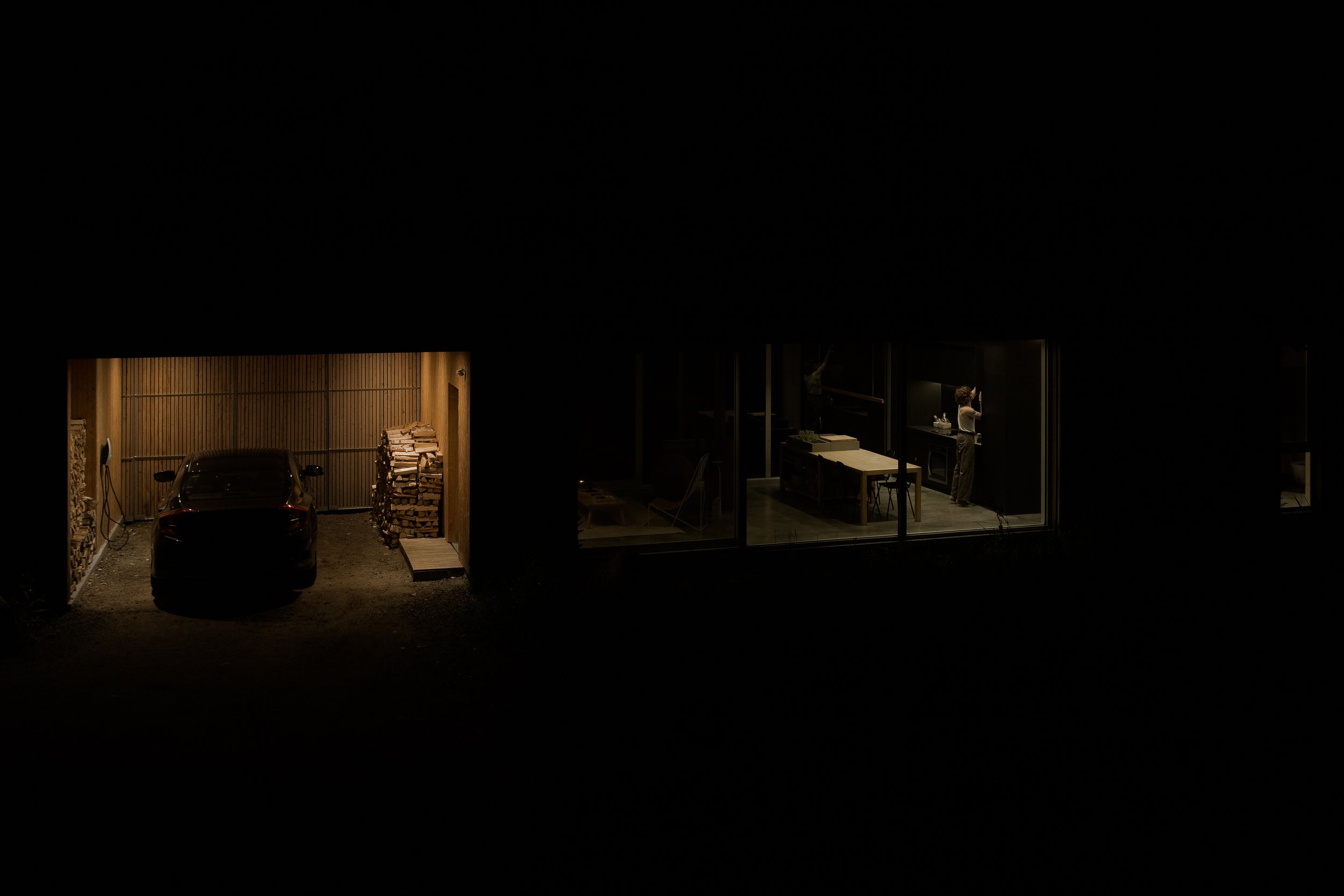Nighttime view of a modern house with a garage on the left, illuminated with warm lighting showing a parked car and stacked firewood, and a brightly lit interior room on the right with a woman cooking in a sleek kitchen.