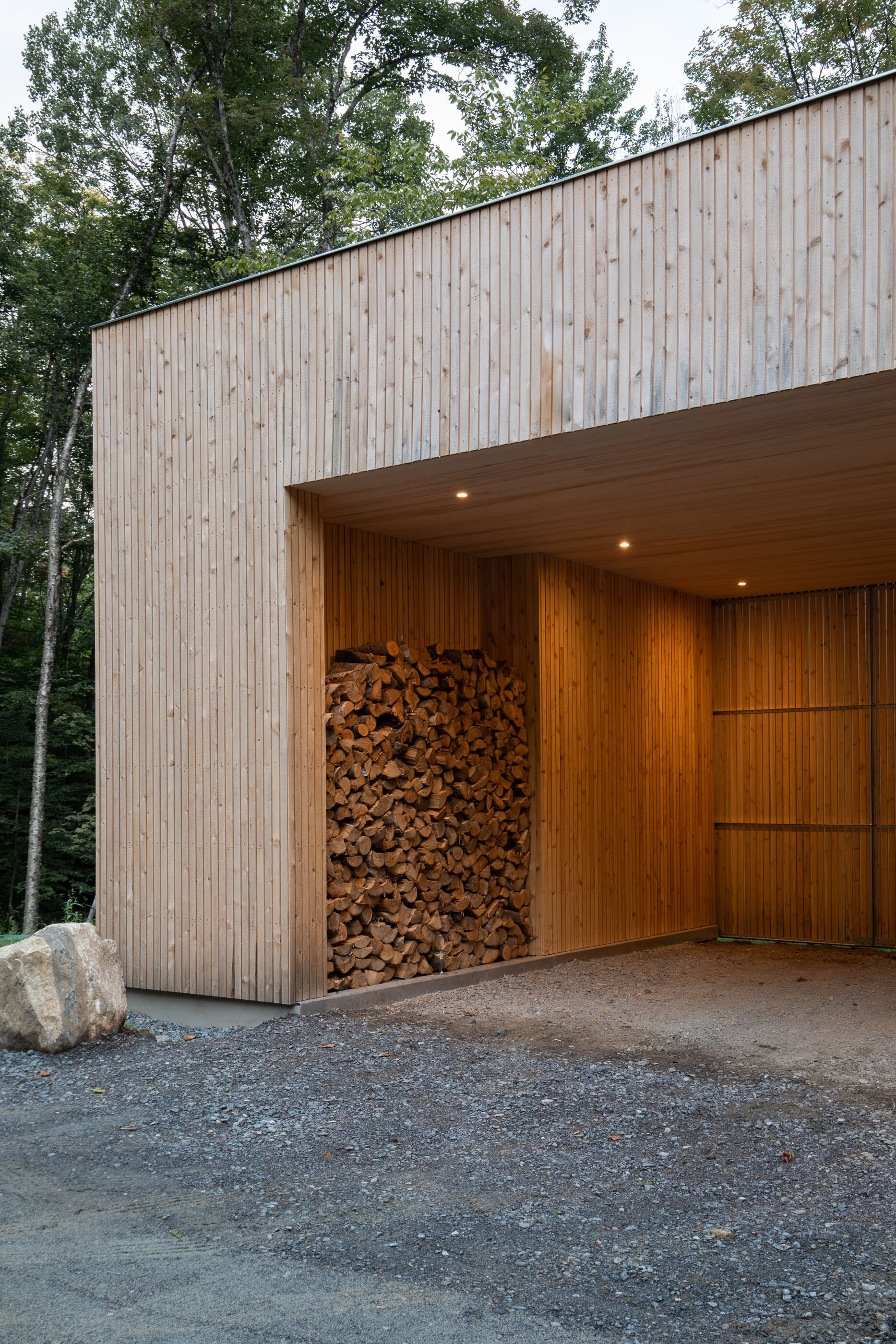 Modern wooden garage with a woodpile stored inside, set against a forested background, featuring a gravel driveway and recessed ceiling lights.