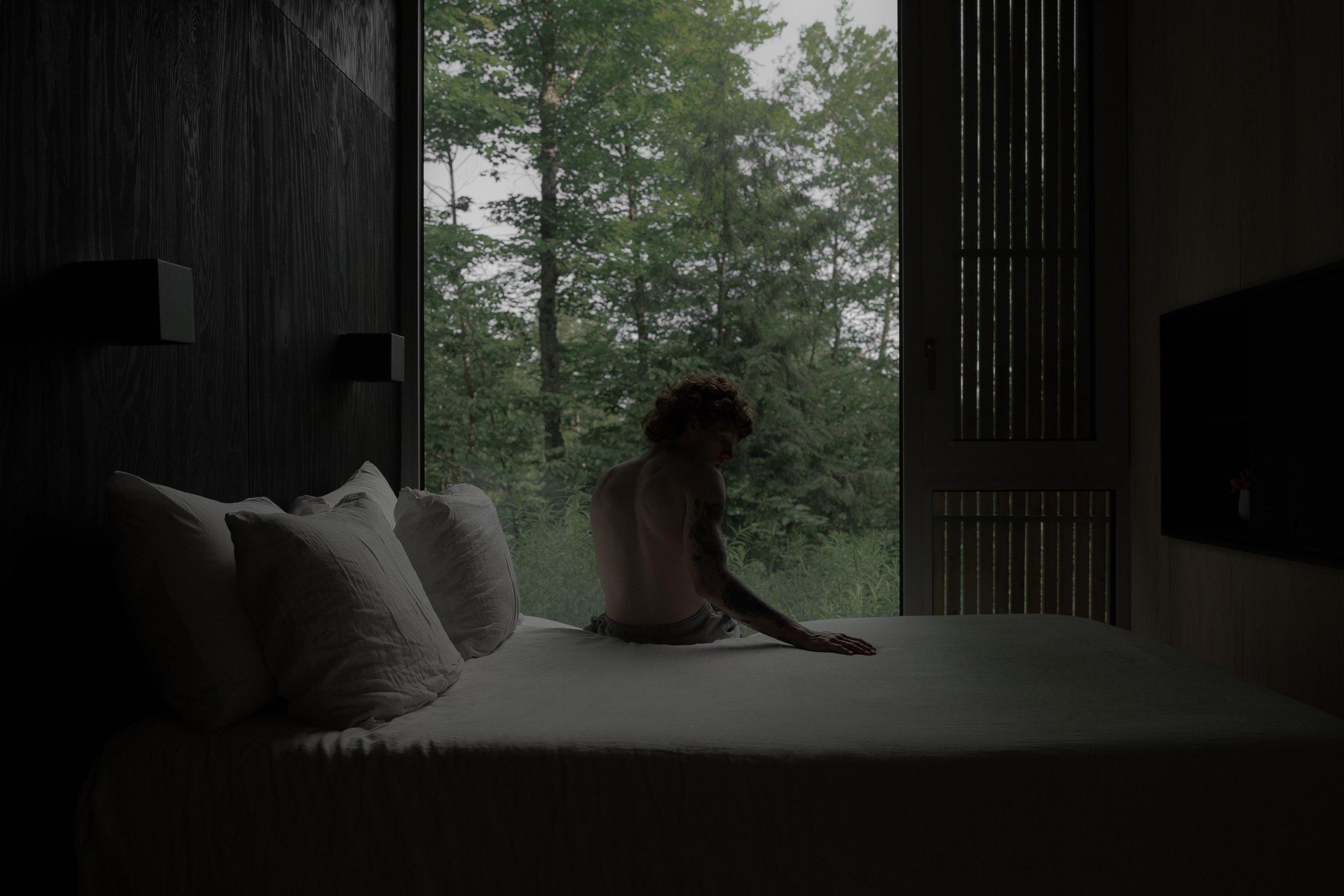 A person sitting on a bed in a dark room with large window showing a forest outside.