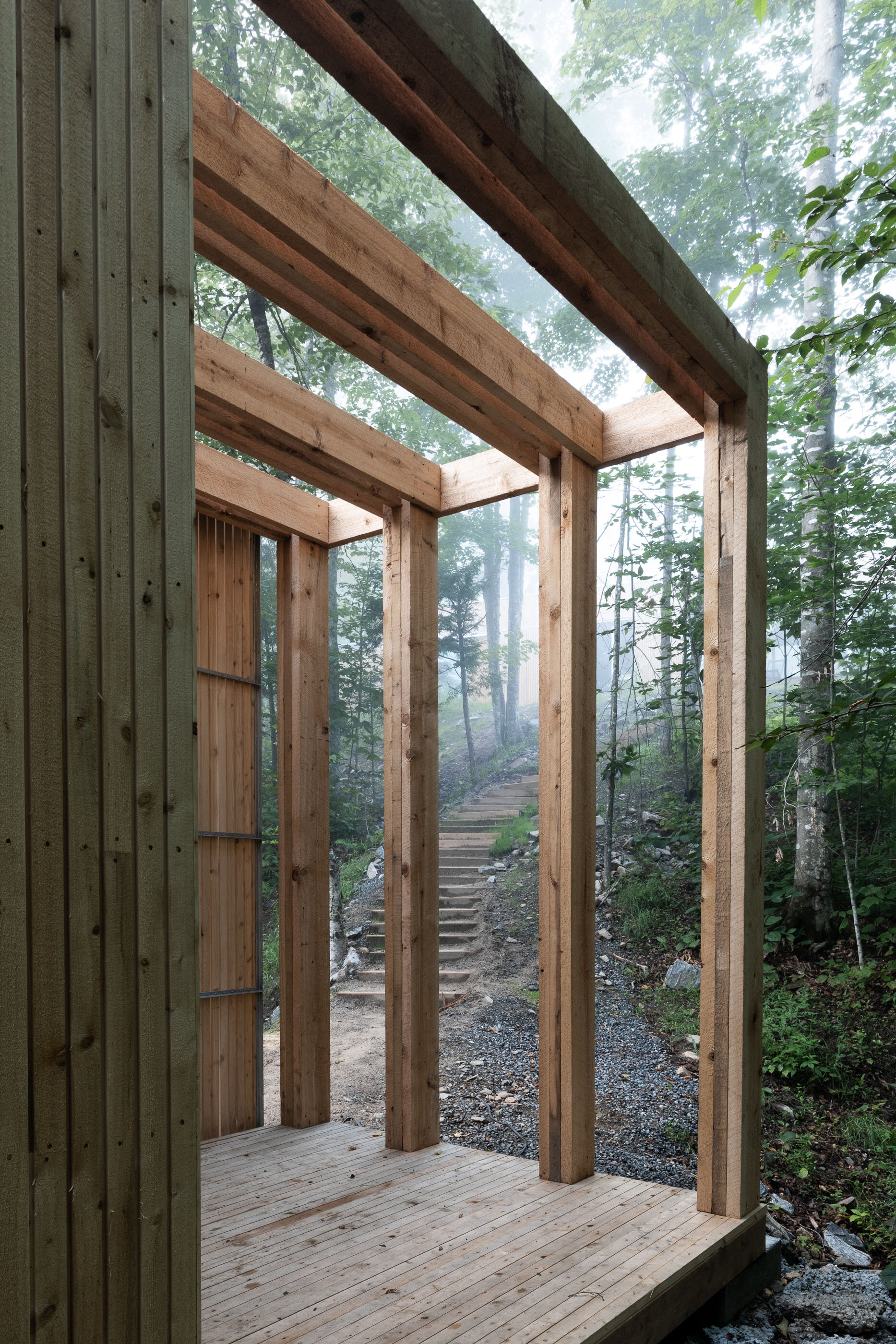 Construction of a wooden outdoor structure, showing a partially built frame with vertical and horizontal beams, overlooking a forested trail with stairs.